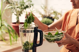 Young housewife putting fresh green spinach leaves into electric blender while preparing healthy smoothie for breakfast