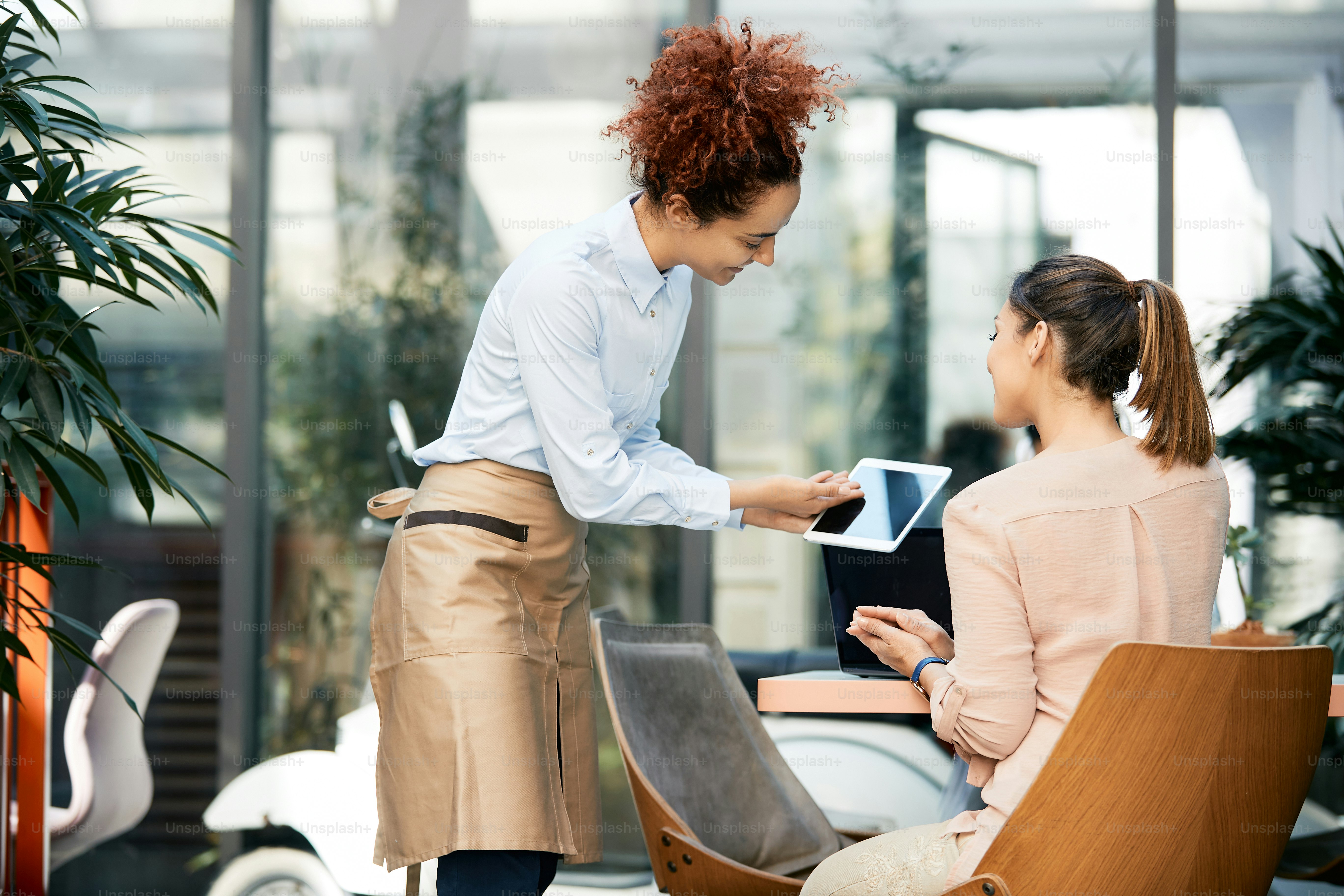 Young waitress using digital tablet while showing menu to her guest in a cafe.
