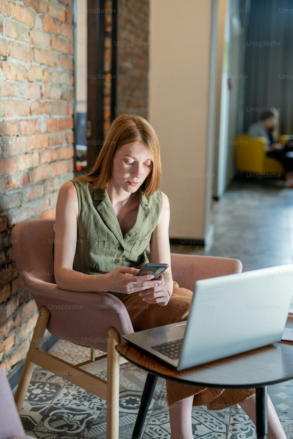 Junge Frau scrollt im Smartphone, während sie im Café oder Büro vor dem Laptop sitzt