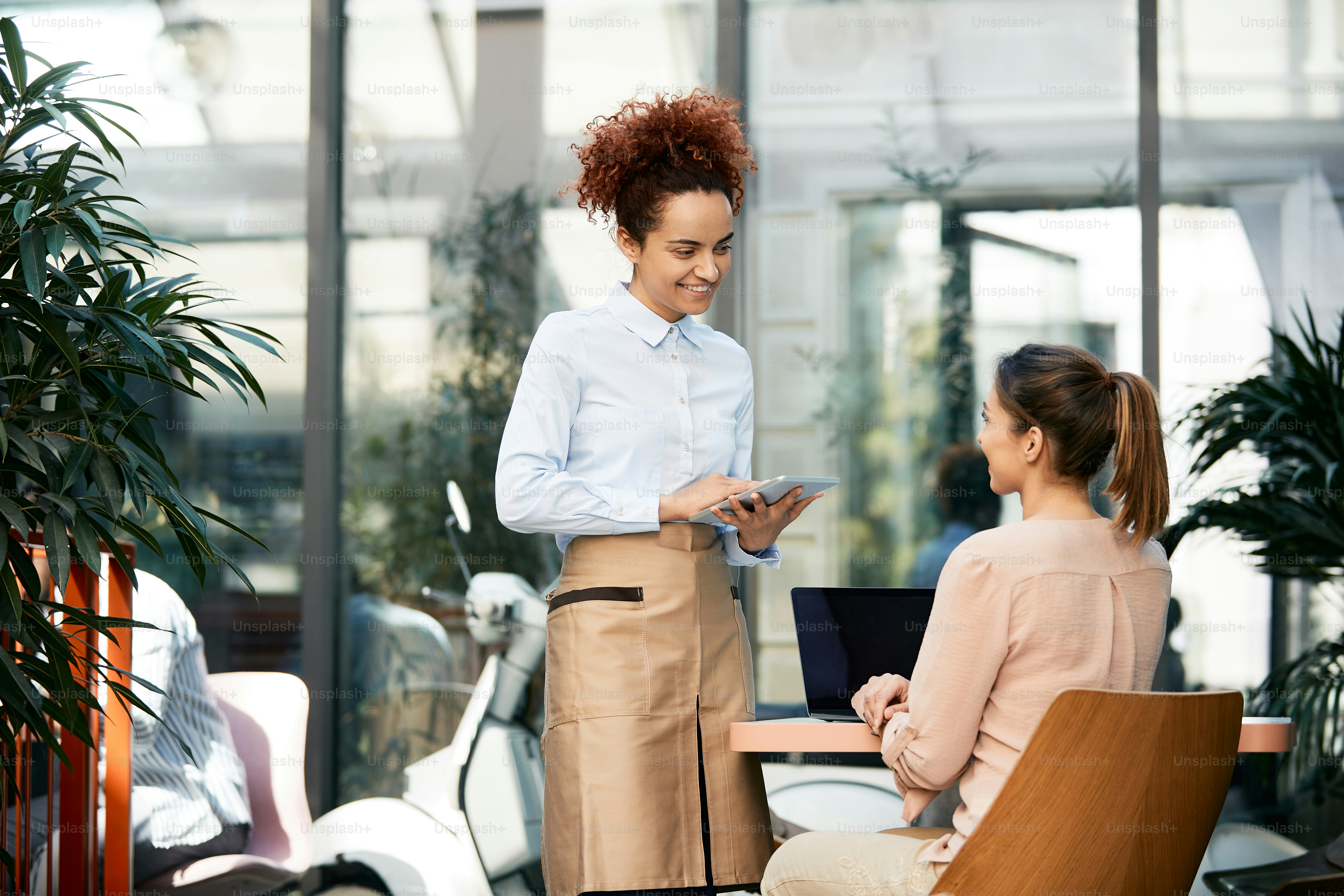 Smiling waitress talking to female guest while using touchpad in a cafe.