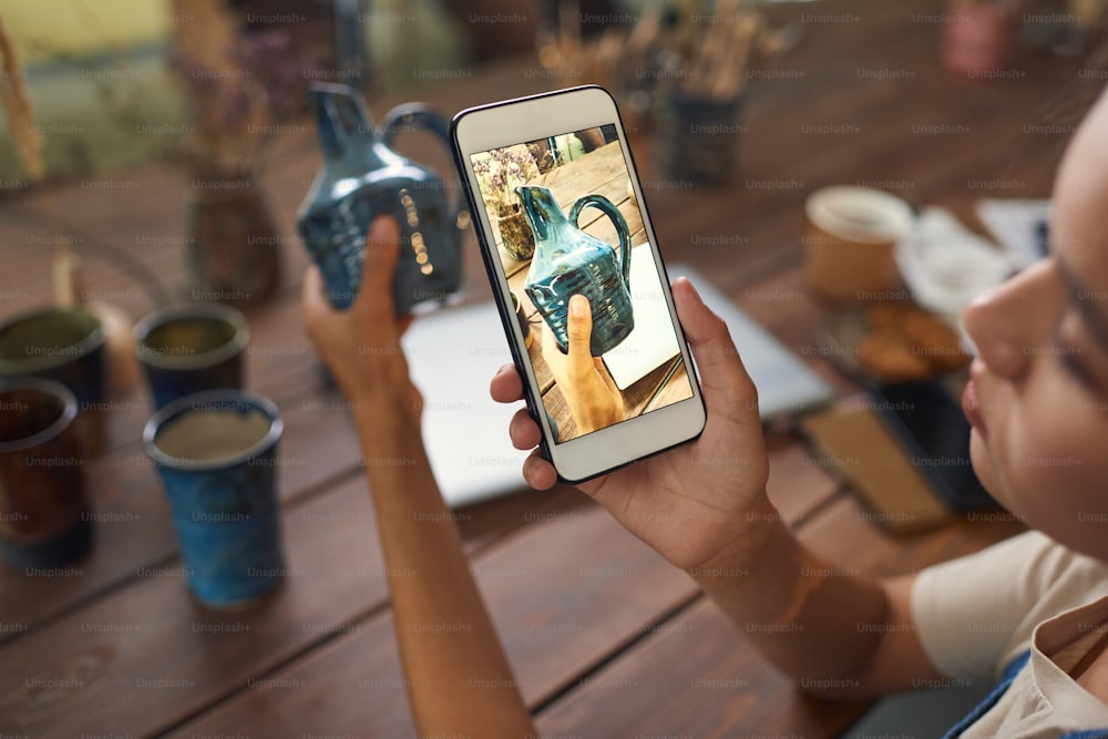 Focus on smartphone screen: potter taking photo of ceramic vase in workshop