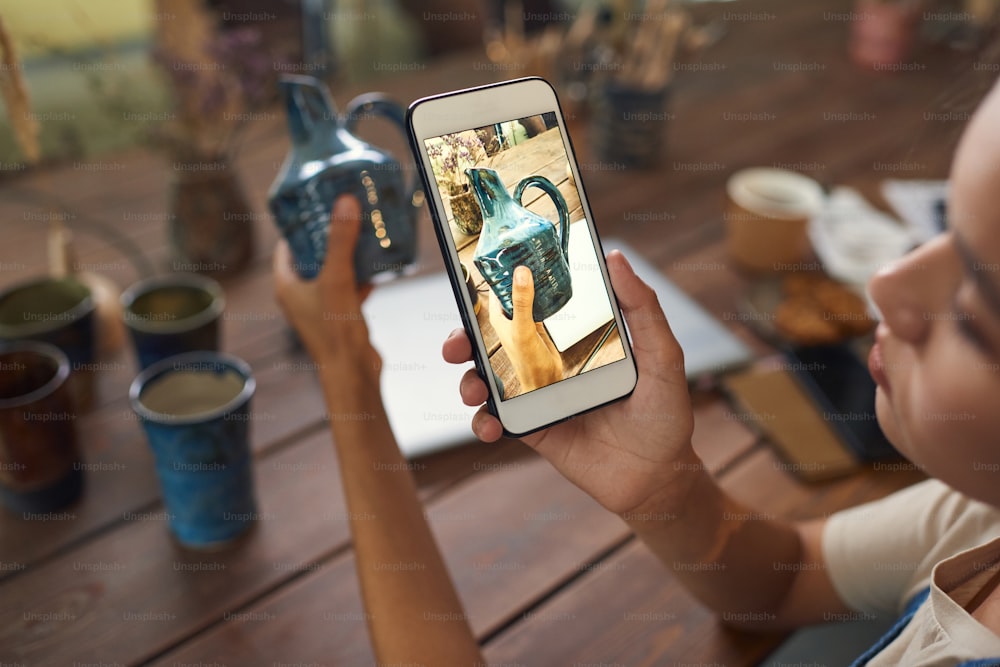 Focus sullo schermo dello smartphone: vasaio che scatta foto di un vaso di ceramica in officina