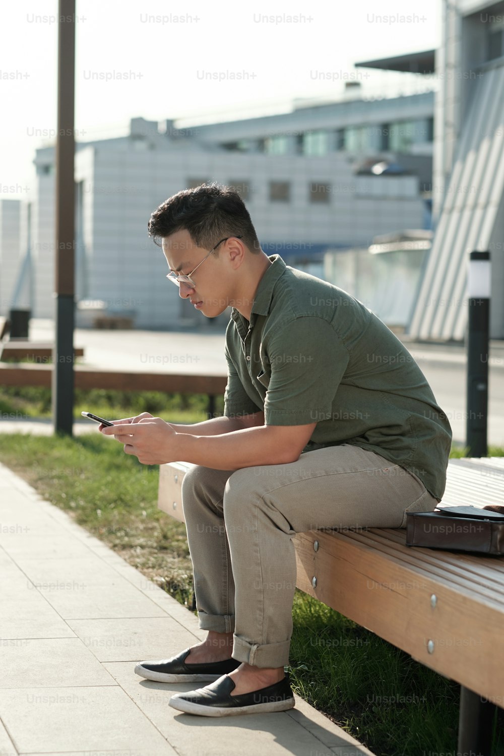 Jovem empresário asiático rolando em smartphone enquanto está sentado no banco em ambiente urbano no dia de verão