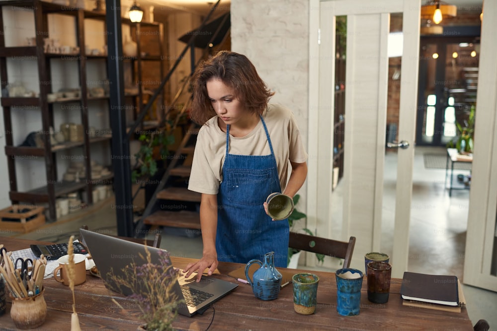 Serious young Caucasian owner of pottery shop in denim apron holding craft mug and using laptop while processing order