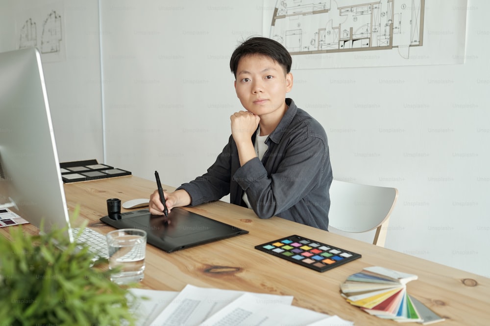 Jeune designer féminin sérieux avec stylet dessin croquis sur l’écran de la tablette graphique tout en étant assis près du lieu de travail