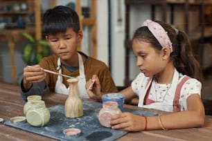 Concentrated interracial kids sitting at wooden table and painting clay vase together in workshop