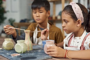 Concentrated multi-ethnic school children sitting at table and using paintbrushes while creating design of vase at class
