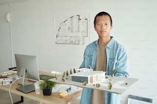 젊은 자신감 있는 건축가는 사무실에서 직장을 배경으로 카메라 앞에 서서 집 레이아웃을 들고 있다