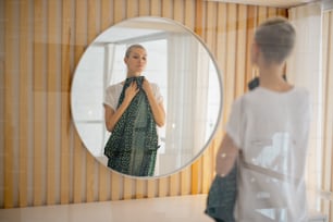 Young caucasian woman looking green dress with dots on herself in mirror. Concept of fashion. Back view of beautiful slim girl. Interior of living room in modern apartment
