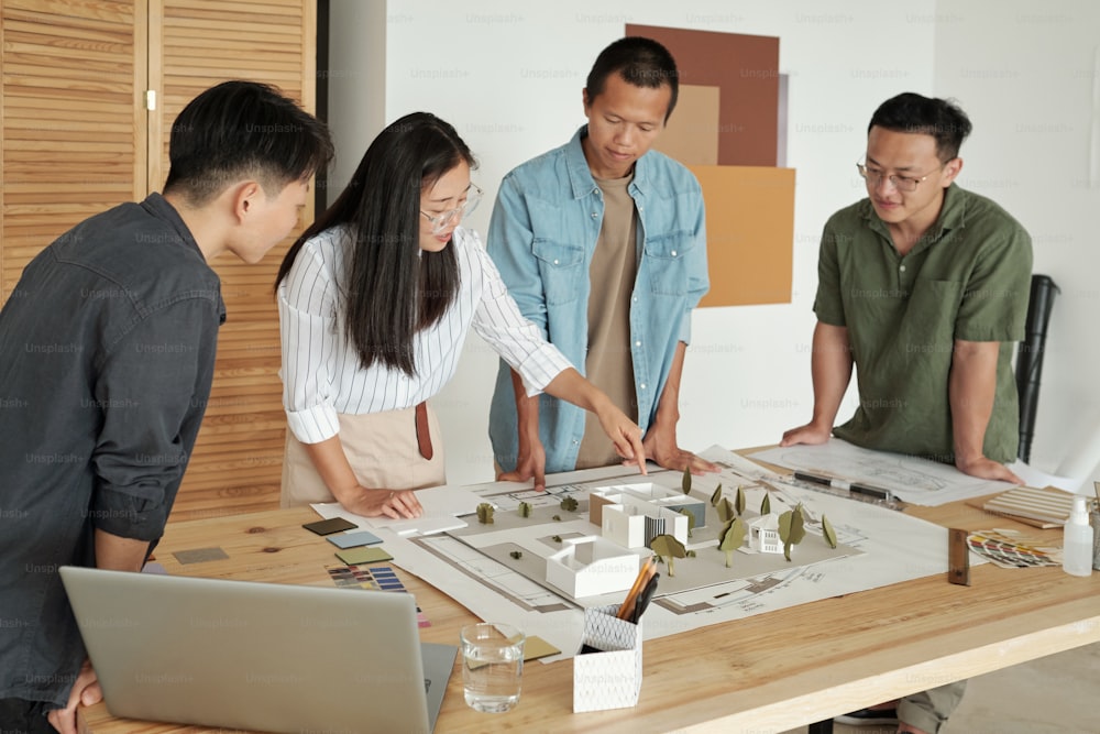 Joven empresaria china señalando el diseño de la casa mientras está de pie entre colegas por lugar de trabajo en la reunión de trabajo