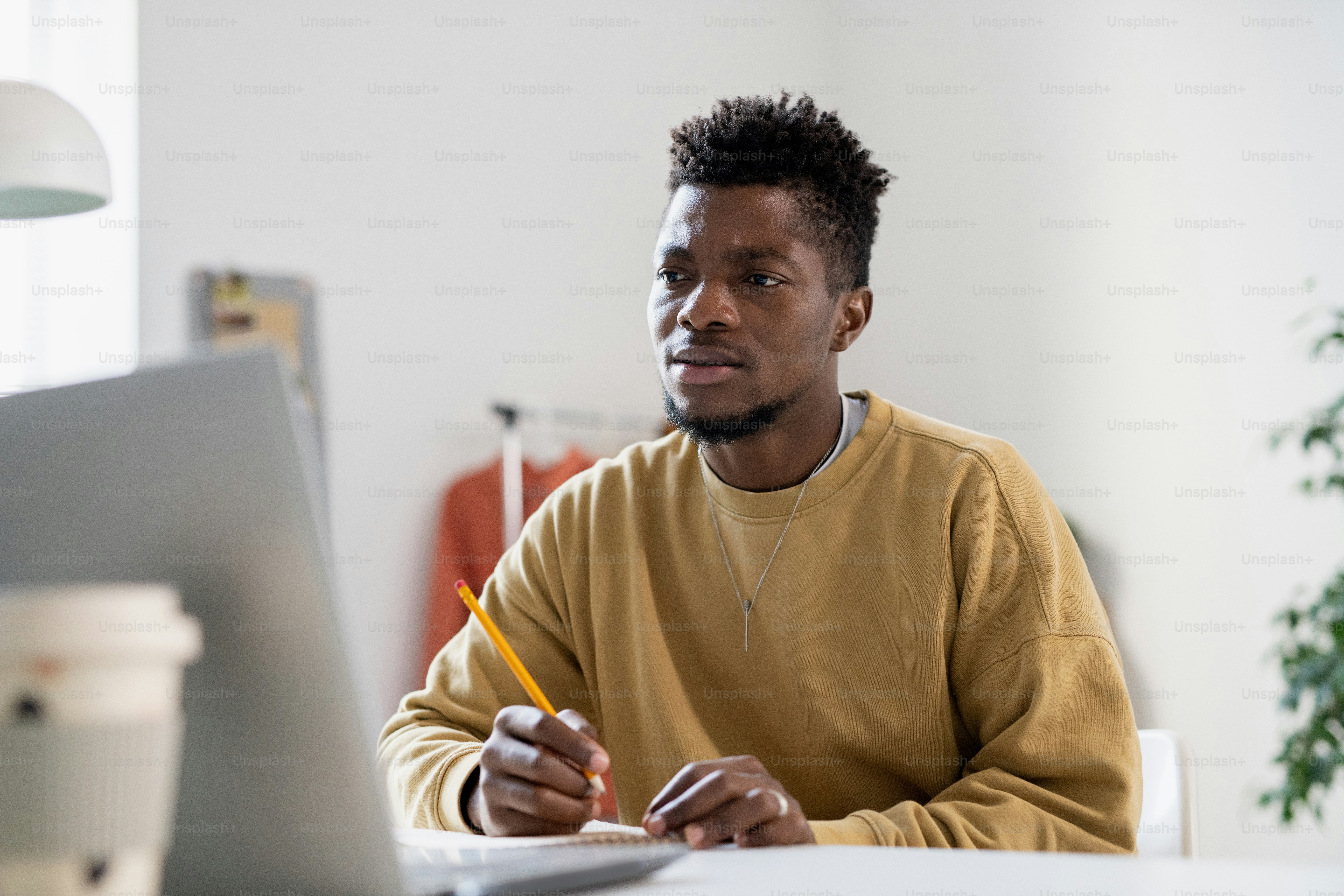 Tense young man with pencil looking at laptop screen while making notes and listening to teacher during online lesson