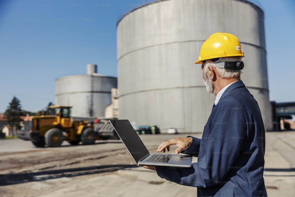 A senior businessman with a helmet on his head holds a laptop and typing on it while visiting his factory. In the background are silos. Factory business and technology.