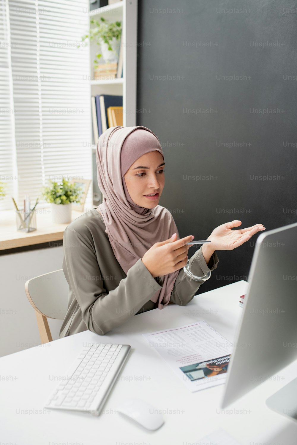 Young confident teacher in hijab speaking to online audience in front of computer monitor while sitting by desk in home environment