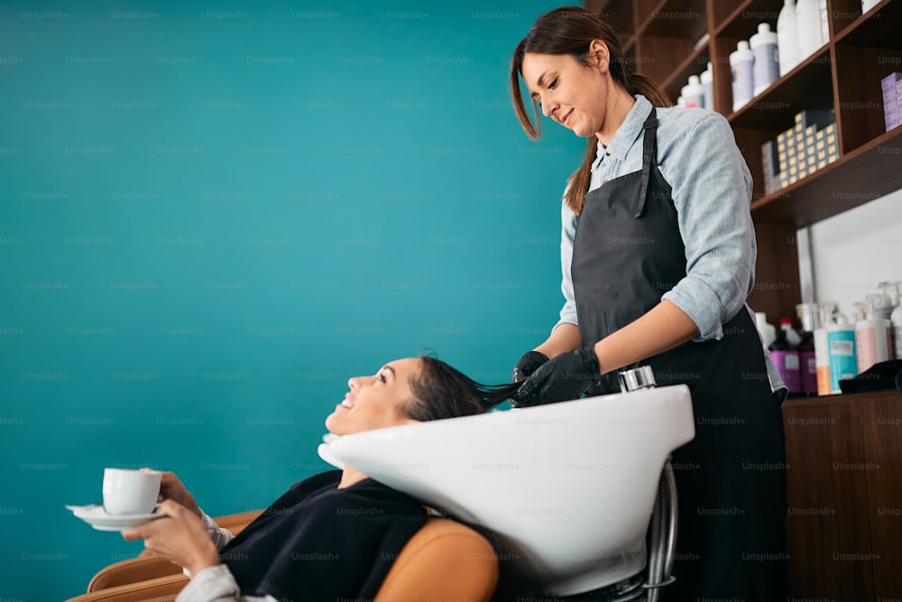 Junge glückliche Frau, die es genießt, eine Tasse Kaffee zu trinken und sich im Friseursalon die Haare waschen zu lassen. Speicherplatz kopieren.