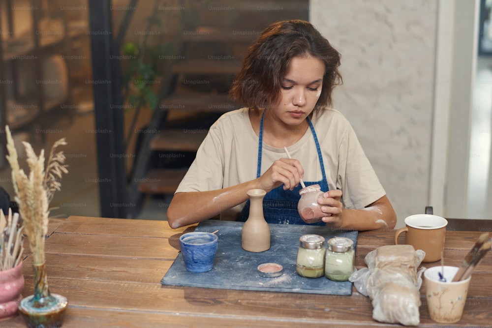 Young serious female painting handmade earthenware while sitting by table and holding one of three jars with paints
