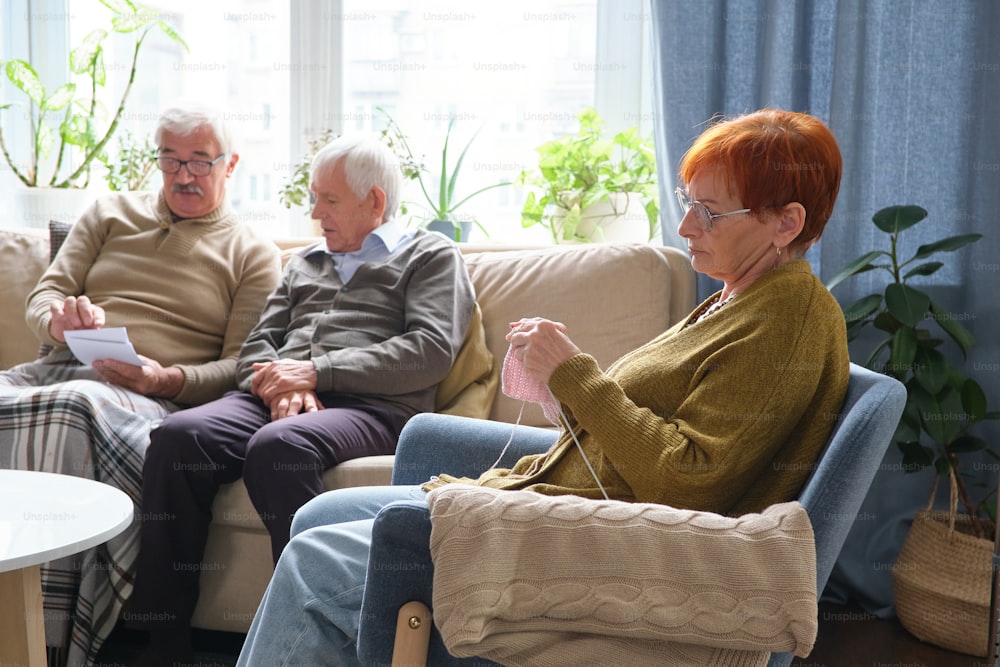 Senior woman sitting in armchair and knitting with two senior men discussing photos together on sofa in the background