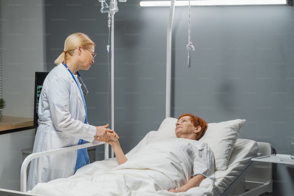 Médico tomado de la mano y hablando con una mujer mayor mientras ella está acostada en la cama después de una operación seria