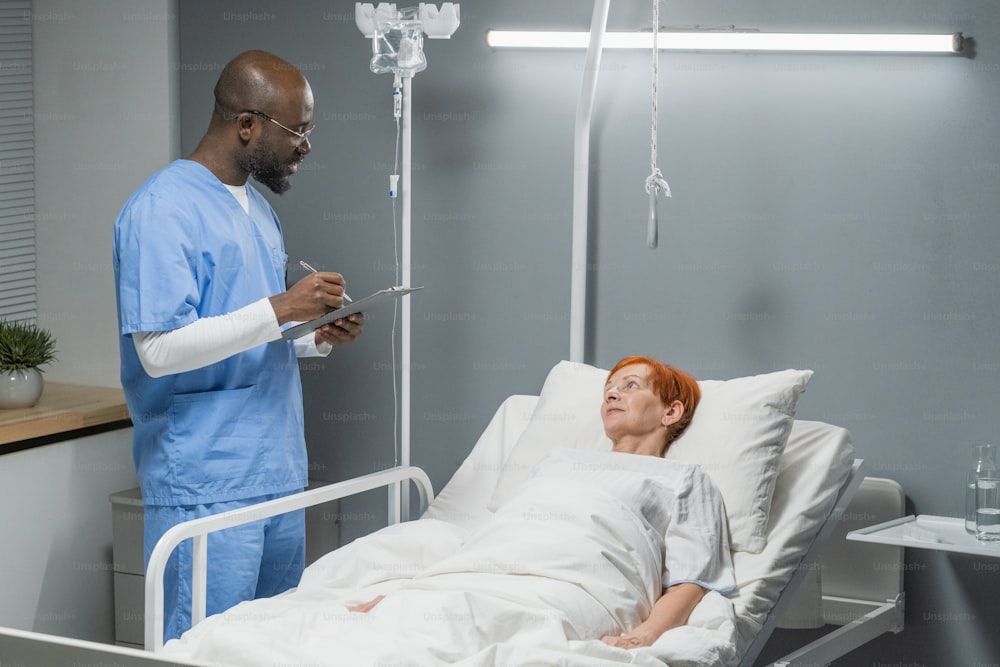 Un médico africano escribe una receta para su paciente mayor mientras ella está acostada en la cama de la sala del hospital