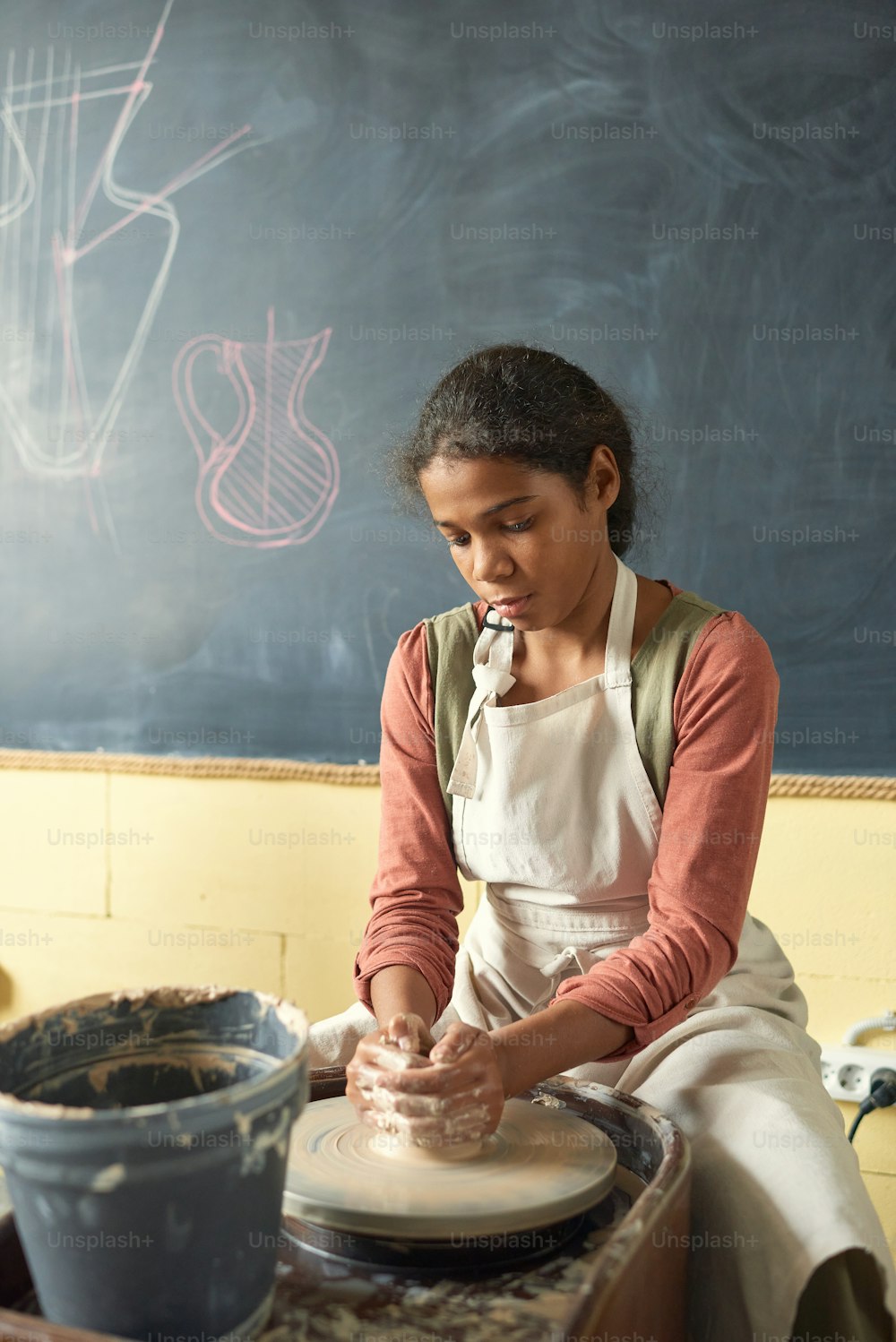 Pretty African schoolgirl in apron sitting by rotating pottery wheel while sculpting jug or pot against blackboard in classroom