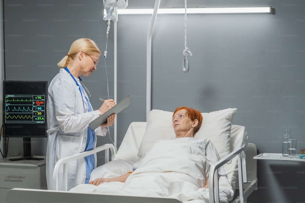 Krankenschwester im weißen Kittel füllt die Krankenkarte des Patienten, der nach der Operation auf der Station auf dem Bett liegt