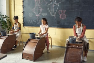 Fila de tres escolares interculturales haciendo vasijas de barro u otra loza mientras están sentados junto a ruedas en la clase de cerámica