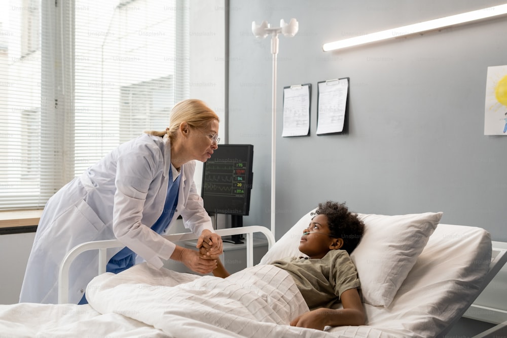 Ärztin im weißen Kittel kümmert sich um kleinen Jungen, der nach der Operation auf der Krankenstation auf dem Bett liegt