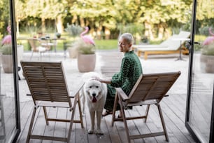 自宅のテラスで木製の椅子に座りながら犬を愛撫する女性。ペットと時間を楽しんでいるヨーロッパの女の子。現代のライフスタイルのコンセプト。女性の友情のアイデア。晴れて暖かい日中