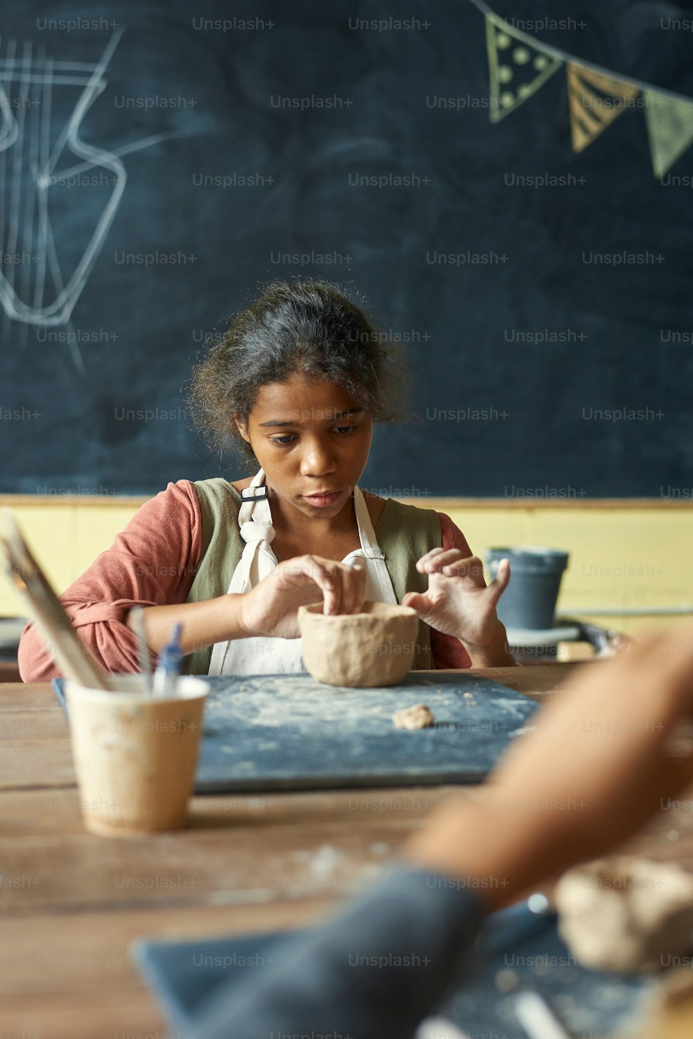 Menina jovem bonito no avental fazendo tigela de barro ou caneca enquanto sentado pela mesa contra o quadro negro na lição de artesanato