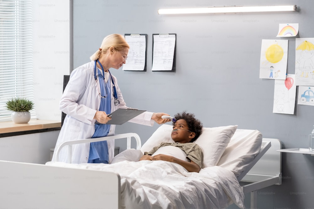 Ärztin im weißen Kittel mit Krankenkarte und Gespräch mit kleinem Jungen, der auf der Krankenstation im Bett liegt