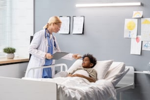 Female doctor in white coat holding medical card and talking to little boy who lying in bed at hospital ward