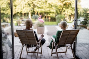 Multiracial couple drinking cocktails while sitting on wooden chairs at home terrace. European girl and black man spending time together. Concept of leisure. Modern domestic lifestyle