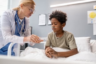 Doctora que hace goteo intravenoso para un niño pequeño que se sienta en la cama durante su tratamiento en el hospital