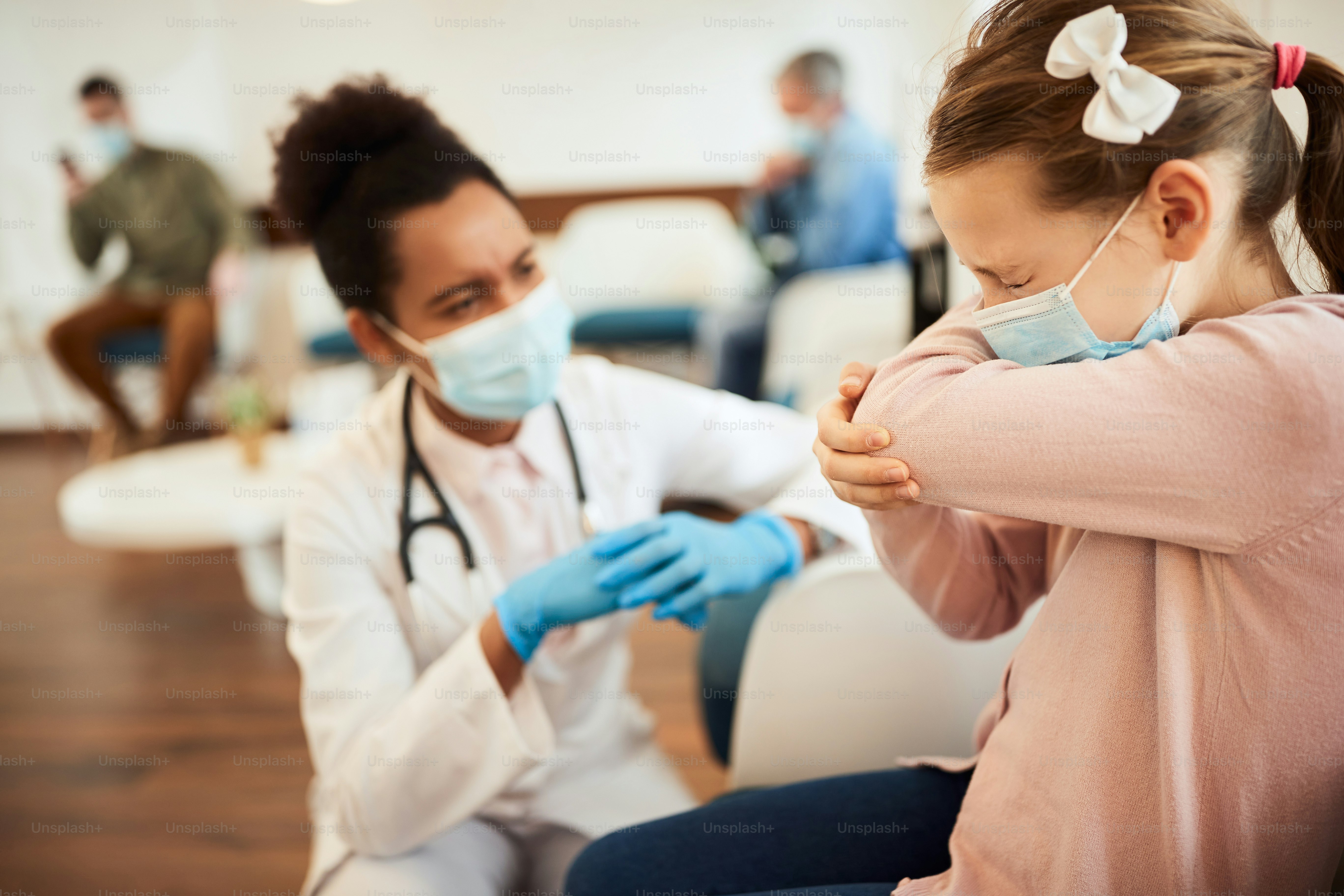 Little girl wearing face mask and coughing into elbow while talking to a doctor in waiting room