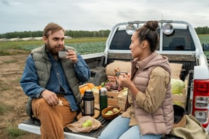 Content young multi-ethnic farmers in warm vests sitting in enclosed cab of pickup truck and chatting while drinking hot tea in field