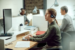 Businesswoman wearing headset while working on a computer in the office. He colleagues are in the background.
