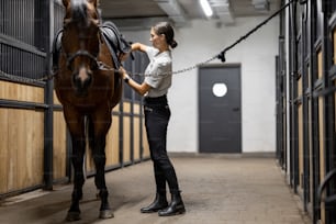 Female horseman puts ammunition on her brown Thoroughbred horse in stable. Concept of animal care. Rural rest and leisure. Idea of green tourism. Young smiling european woman wearing uniform