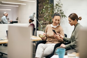 Happy Asian entrepreneur showing something on mobile phone to her female colleague during their lunch break in the office.