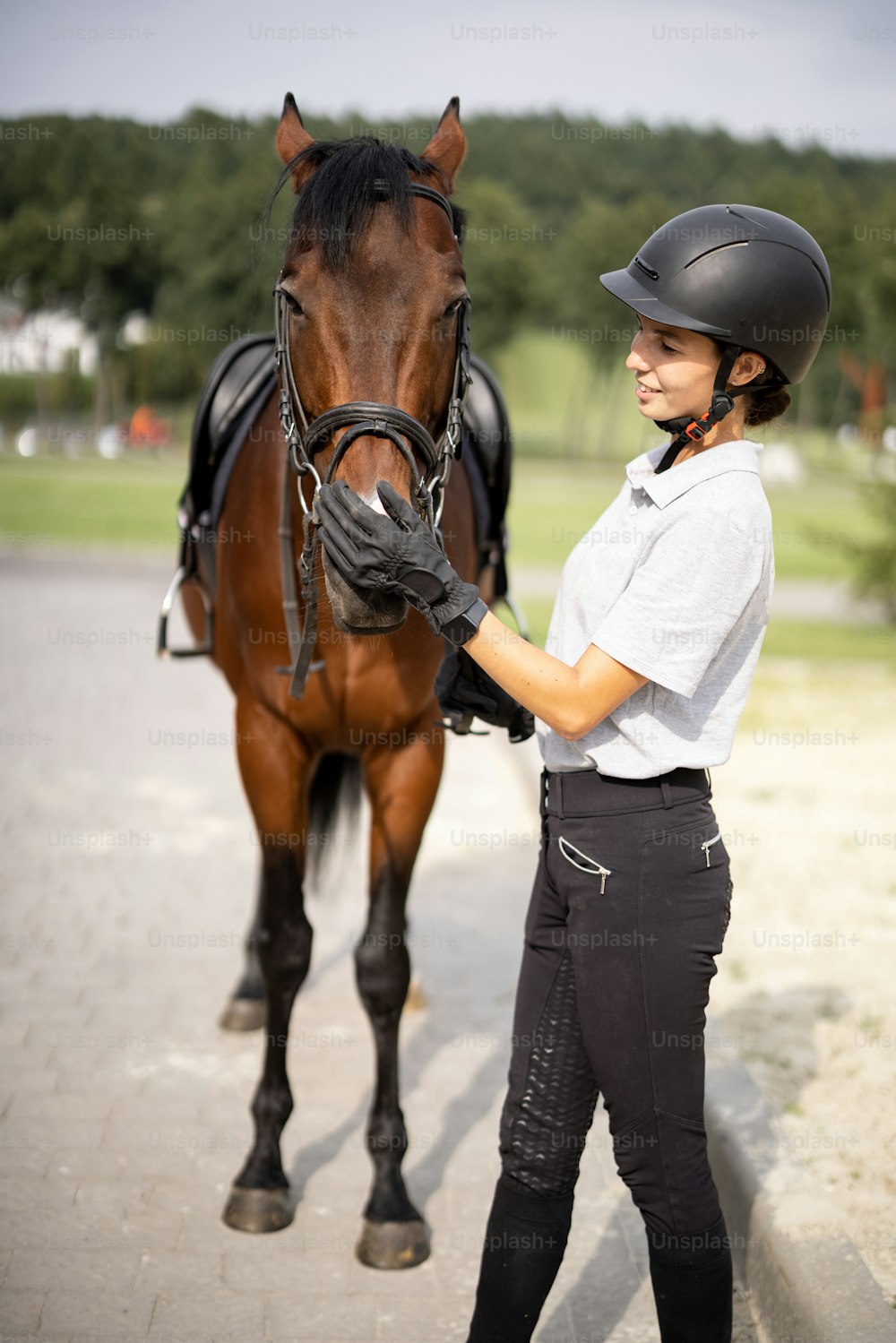 Female horseman with her brown Thoroughbred horse near stable outdoors. Concept of animal care. Rural rest and leisure. Idea of green tourism. Young european woman wearing helmet and uniform