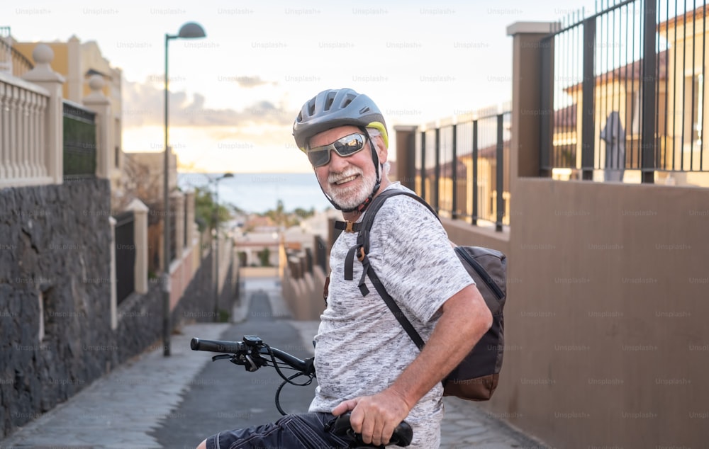 スポーツヘルメットとサングラスをかけた笑顔の年配の男性が、夕暮れ時に電動自転車で自転車をサイクリングするカメラを見ています。背景に水面の地平線