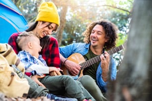 Happy family camping in the forest playing guitar and singing together - Mother, father and son having fun trekking in the nature sitting in front of the tent - Family, nature and trekking concept