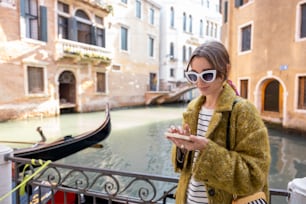 Young woman using phone while walking near beautiful water canals in Venice. Concept of vacations in Italy. Caucasian stylish woman wearing coat sunglasses and colorful shawl