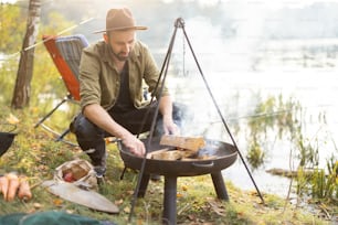 自然の中で料理をするためにグリルで火を焚く白人男性。自然の中でのレジャー、週末、趣味、休暇のコンセプト。秋の日に湖や川の海岸で休んで釣りをする男性