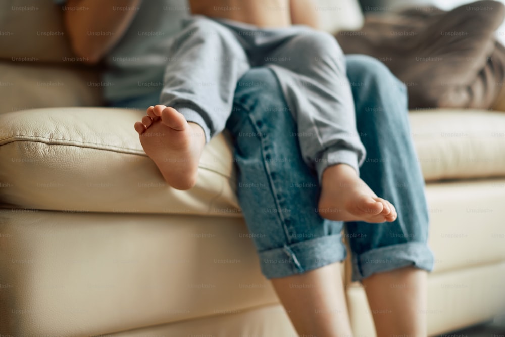 Close-up of barefoot boy sitting on mother's lap while being at home together.