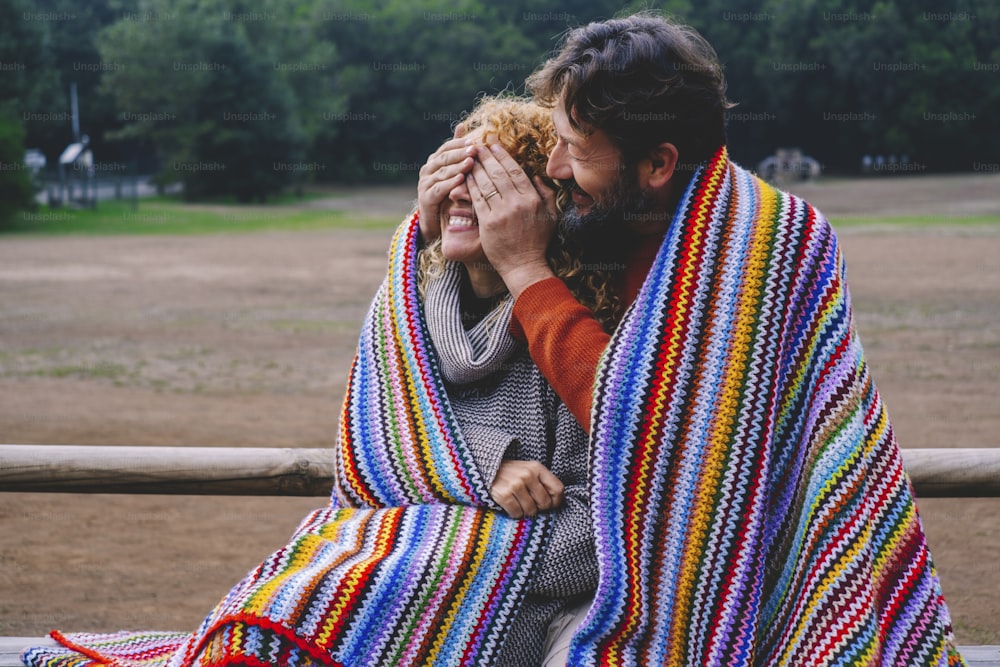 Happy couple enjoy and play under a colorful blanket outdoor at the park. Man close woman eyes to do a surprise and have fun together in leisure activity
