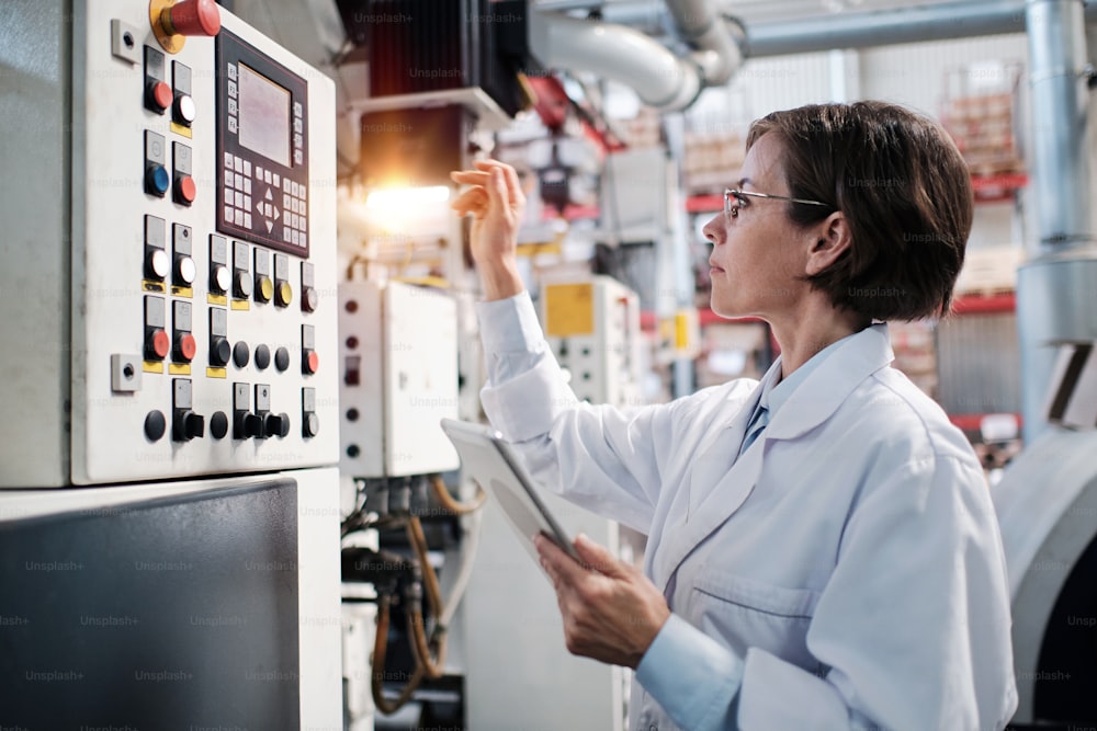 Portrait of female factory worker wearing glasses and white robe, pressing machine buttons, using tablet for quality control and logistic purposes at polymer plastic manufacturing