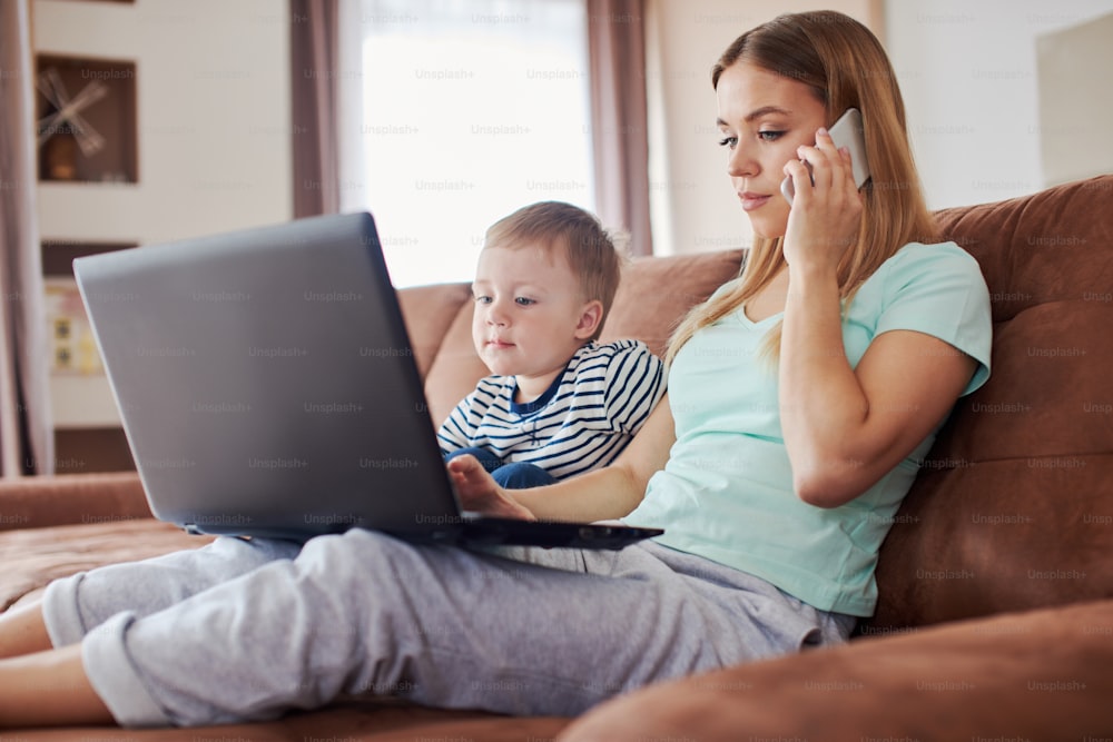 Giovane madre caucasica con figlio piccolo sta usando il computer portatile e lo smartphone mentre è seduta sul divano di casa, indossando il pigiama
