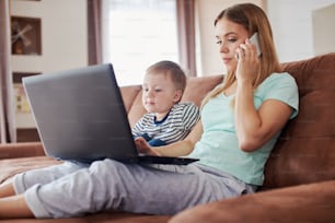 Young caucasian mother with toddler son is using laptop and smart phone while sitting on the couch at home, wearing pyjamas