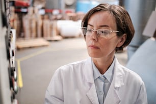 Portrait of female factory worker wearing glasses and white robe, using tablet for quality control and logistic purposes at polymer plastic manufacturing