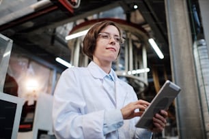 Portrait of female factory worker wearing glasses and white robe, using tablet for quality control and logistic purposes at polymer plastic manufacturing