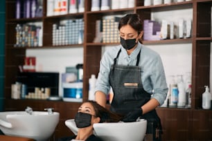 La parrucchiera e la sua cliente indossano maschere protettive per il viso durante il lavaggio dei capelli in salone.